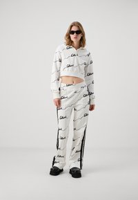 Fehér cropped kapucnis pulóver és hozzá illő nadrág fekete feliratos logóval, puha anyagból készült, laza szabással és kontrasztos oldalsávokkal. Fekete, vastag talpú cipő.