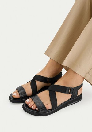 Nohy v černých sandálech s otevřenou špičkou, širokými elastickými a koženými pásky, v kombinaci s béžovými zkrácenými kalhotami na světlém pozadí.