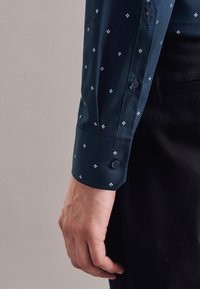 Männliche Hand liegt neben dem Körper, trägt einen dunkelblauen Hemdärmel mit kleinem weißem Diamantmuster und schwarze Hose.