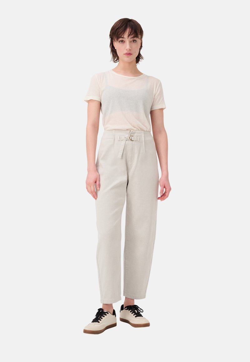 T-shirt beige clair à manches courtes, associé à un pantalon gris clair taille haute avec une ceinture et des détails en anneau, et des baskets simples noires et crème.