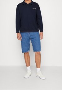 Marinblå långärmad polo med krage och logotyp, tillsammans med knälånga blå denimshorts. Vita sneakers och strumpor i ankellängd fulländar looken.