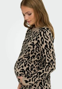 Femme enceinte portant un pull beige et noir à motif animal, tenant doucement son ventre, le regard baissé avec une expression douce.