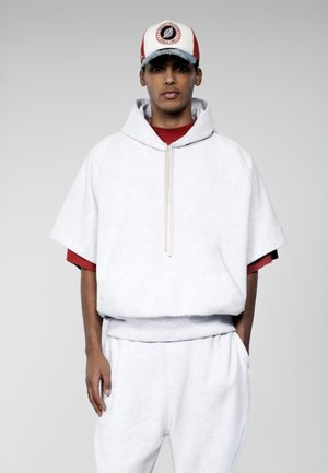 Jeune homme portant un sweat à capuche blanc à manches courtes par-dessus un t-shirt rouge, un pantalon de survêtement blanc et une casquette de baseball blanche et rouge, debout avec une main dans la poche.