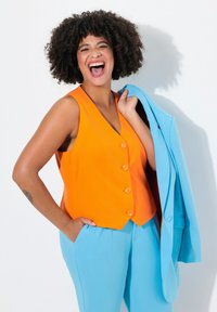 Oranje mouwloze vest met vijf knopen, gecombineerd met lichtblauwe op maat gemaakte broek en een bijpassende jas die in één hand wordt vastgehouden tegen een witte achtergrond.