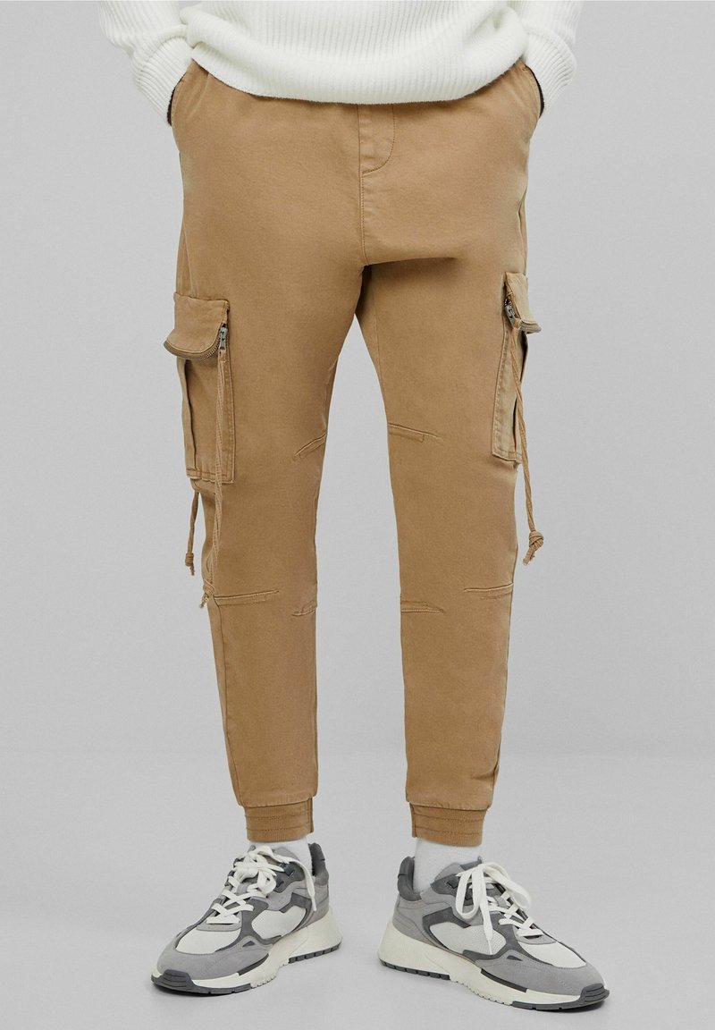 Tan cargo byxor med slitna detaljer, dragkedjefickor på sidorna, smal passform och uppvikkta anklar. Matchat med grå och vita sneakers.