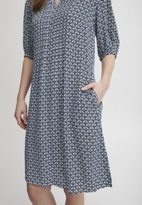 Modrá a bílá šaty s vzorem, krátkými volánovými rukávy, výstřihem do V a bočními kapsami. Mají geometrický design a volný střih.