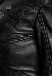 Close-up van een zwart leren jack met een zak met knoop en gedetailleerde stiksels op het gestructureerde materiaal.