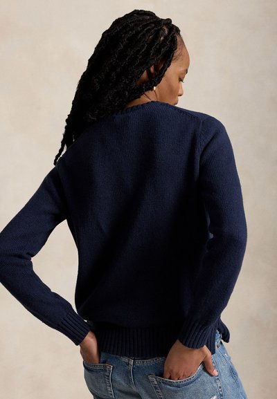 Moteris su ilgais susukti plaukais, dėvinti tamsiai mėlyną megztinį ir šviesiai mėlynas džinsines kelnes, stovinti su rankomis užpakalinėse kišenėse.