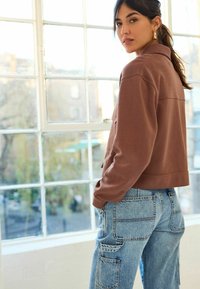 Braune cropped Jacke mit spitzem Kragen, kombiniert mit hellblauen Cargo-Jeans, die mehrere Taschen und eine Denim-Textur aufweisen. Fenster im Hintergrund.