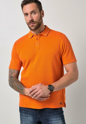 Mężczyzna z krótką brodą, ubrany w pomarańczową koszulkę polo z krótkim rękawem, ciemne dżinsy, zegarek i tatuaż na lewym przedramieniu, stojący na jednolitym tle.