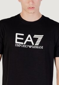 Camiseta de algodón negra con un logo blanco que presenta el texto "EA7" y "EMPORIO ARMANI", cuello redondo y mangas cortas.