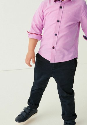 Enfant portant une chemise rose à boutons avec les manches retroussées, un pantalon noir à revers et des baskets noires à scratch.