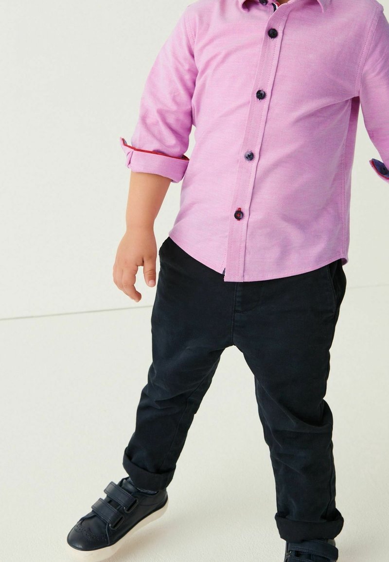 Enfant portant une chemise rose à boutons avec les manches retroussées, un pantalon noir à revers et des baskets noires à scratch.