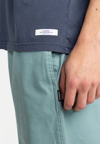 Dunkelblaues T-Shirt mit kurzen Ärmeln und einem weißen Etikett mit der Aufschrift "ELEMENT SKATEBOARD CO." sowie hellgrünen Shorts mit einem schwarzen Etikettendetail.