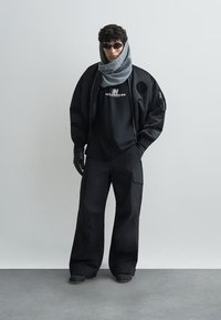 Sort bomberjakke over en sort T-shirt, brede sorte bukser, grå tørklæde, sorte handsker og solbriller; har et logo på T-shirten.