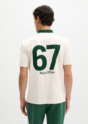 Muškarac u bijeloj majici kratkih rukava s zelenim ovratnikom i velikim zelenim brojem "67" te tekstom "Marc O’Polo" na leđima, u kombinaciji sa zelenim hlačama.