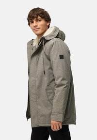 Gray outerwear jacket with a hood, featuring a sherpa-lined collar, side pockets, and a small branded patch on the sleeve.