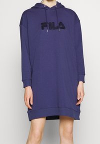 Kvinna som bär en översized navy blue Fila hoodieklänning med långa ärmar och logotyp framtill, stående mot en enkel grå bakgrund.