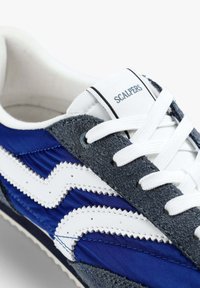 Zapatillas que combinan nylon azul y ante gris, con acentos y cordones blancos en forma de zigzag. Logotipo "SCALPERS" en la lengüeta.