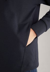 Felpa navy in tessuto morbido e texturizzato; presenta una tasca laterale e polsini aderenti. Il design enfatizza il comfort e l'abbigliamento informale.