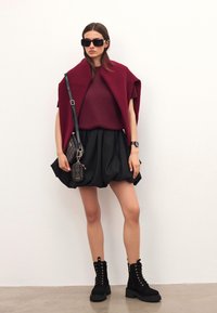 Burgundské pletené tričko a plášť spárované s černou plisovanou sukní. Černé šněrovací kotníkové boty, velké sluneční brýle a vzorovaná crossbody taška.