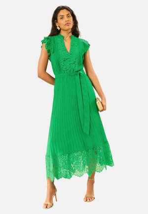 Frau trägt ein grünes Midikleid mit Spitzen-Details, kurzen Rüschenärmeln, einem Taillenband, hält eine goldene Clutch und trägt goldene High-Heel-Sandalen.