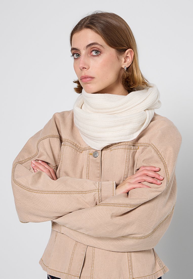 Junge Frau mit hellbraunen Haaren, die eine beigefarbene Jacke und einen dicken weißen Strickschal trägt, mit verschränkten Armen und einem leicht seitlichen Blick.
