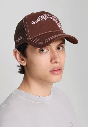 Młody mężczyzna o jasnej karnacji, noszący brązową czapkę Ed Hardy i jasnoszarą bluzę, patrzący lekko na bok na jednolitym tle.