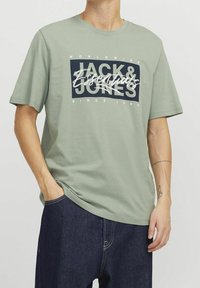 Camiseta de algodón verde claro con un logo gráfico audaz en azul marino y blanco. Manga corta y ajuste relajado, combinada con vaqueros de mezclilla oscuros.