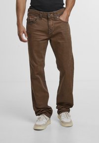Jeans de mezclilla marrones con un corte relajado, que presentan costuras en contraste, cinco bolsillos y cierre de botón y cremallera. Llevados con zapatillas de color claro.