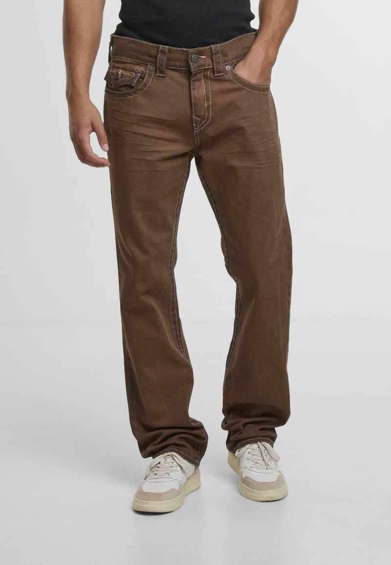 Jeans in denim marrone con una vestibilità comoda, caratterizzati da cuciture a contrasto, cinque tasche e chiusura con bottone e zip. Indossati con sneakers di colore chiaro.