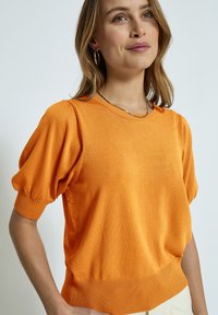Oranžinė megzta džemperis su trumpomis pūkuotomis rankovėmis ir raukšlėtu apatiniu kraštu. Lygus paviršius su apvaliu iškirpimu ir subtiliomis detalėmis ant apykaklės.