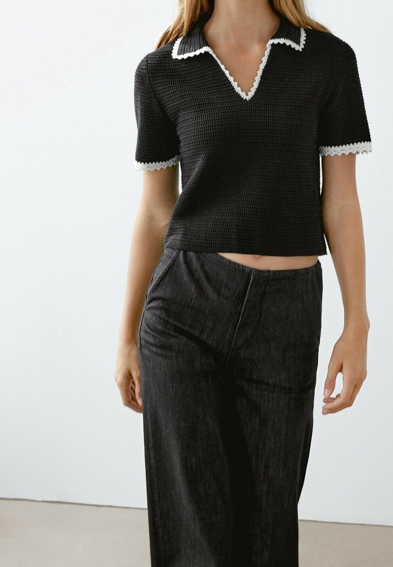 Polo noir en maille avec manches courtes, col et poignets blancs festonnés, coupe courte. Porté avec un pantalon sombre taille haute.