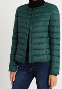 Veste matelassée en vert foncé, avec une conception matelassée, un col rond et des fermetures à pression. Portée avec un t-shirt à col roulé noir et un jean.