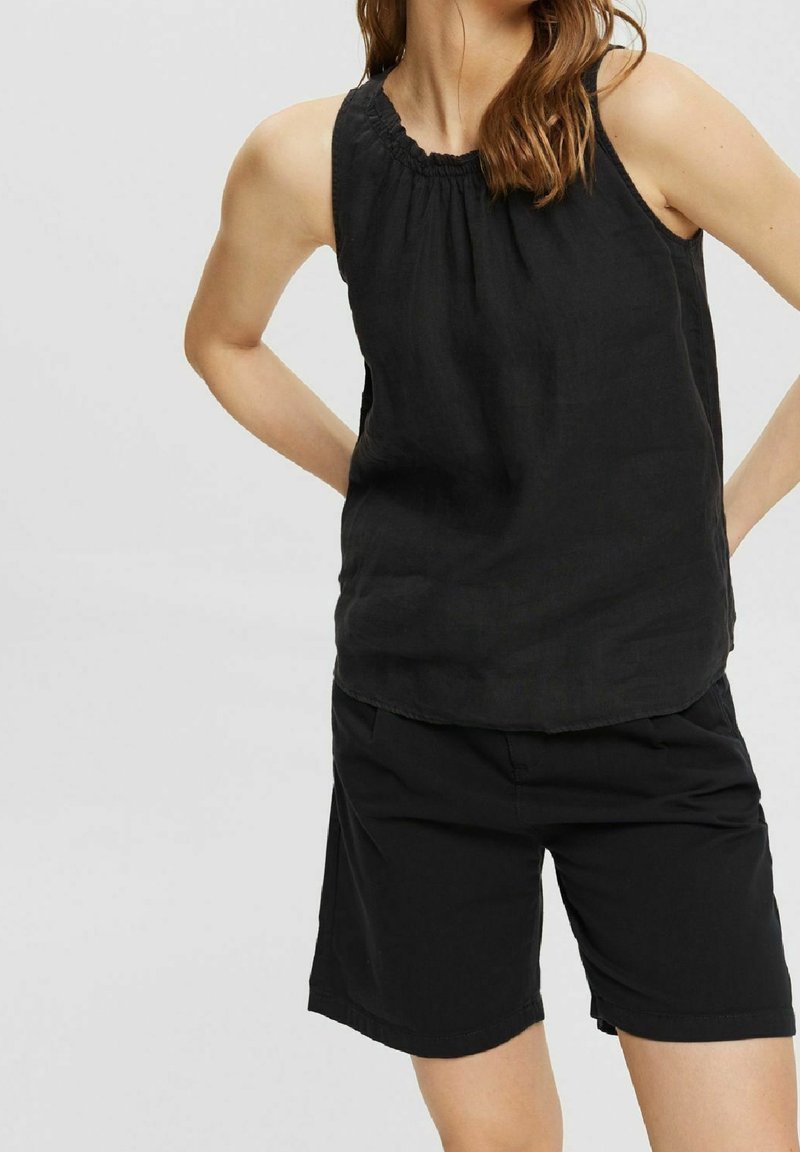 Femme portant un haut noir sans manches avec un col froncé et un short noir jusqu'au genou, debout les mains dans le dos.