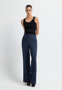 Vrouw staand tegen een effen lichte achtergrond, gekleed in een zwarte mouwloze top, blauwe gestreepte wijde pantalon en zwarte schoenen.