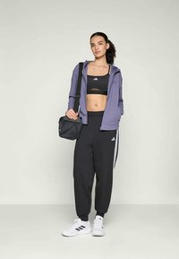 Top cropped atlético preto e calças de treino com riscas brancas da Adidas, combinados com um hoodie roxo com fecho éclair e ténis brancos. Uma bolsa pequena também é visível.