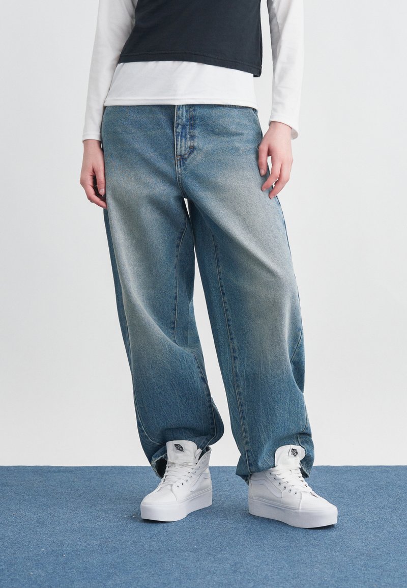 Άτομο ντυμένο με φαρδιά ξεθωριασμένα μπλε τζιν, λευκά ψηλά αθλητικά παπούτσια, λευκό μακρυμάνικο πουκάμισο και μαύρο αμάνικο τοπ σε μπλε χαλί.