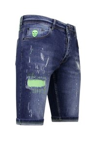 Jeanshort, donkerblauw met versleten details, patches en een groene schedelaccent op de achterzak. Rafelige zoom en gestructureerde stof.