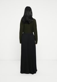 Kvinna med långt svart hår som bär en mörkgrön tröja och en lång svart plisserad kjol, står med ansiktet mot en vit vägg.