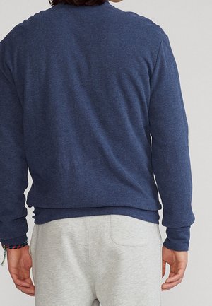 Person trägt ein dunkelblaues Langarmshirt und hellgraue Hose, von hinten gezeigt mit einem mehrfarbigen Armband am linken Handgelenk.