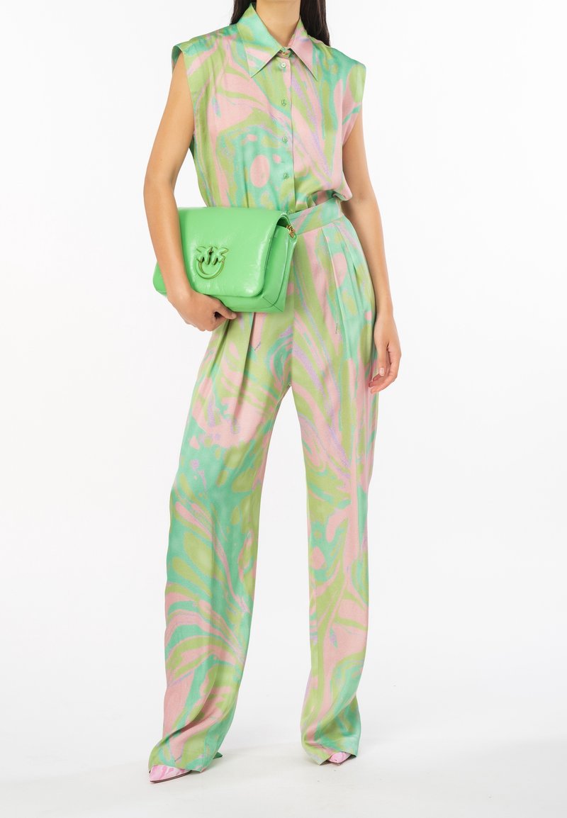Femme portant un chemisier sans manches à motifs vert pastel et rose et un pantalon à jambes larges, tenant un sac à main vert vif avec un détail du logo.