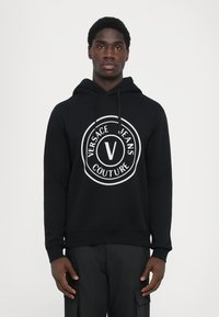 Mustallaneest valmistatud kapuuts, millel on suur valge ümmargune logo, millel on tekst "VERSACE JEANS COUTURE" ja keskel "V". Kapuutsi kinnitusnöör, soonikuga mansetid.