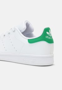 Bílé nízké tenisky Adidas Stan Smith s zeleným pásem na patě, perforovanými třemi pruhy po stranách a bílými tkaničkami na jednobarevném pozadí.