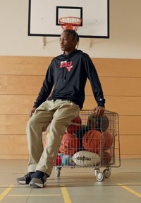 Joueur de basketball portant un sweat à capuche noir et un pantalon cargo beige, assis sur un chariot de courses en fil rempli de ballons de basketball colorés et de ballons de sport.