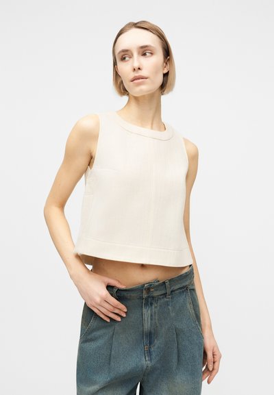 Femme aux cheveux blonds courts portant un haut court blanc sans manches et un jean taille haute bleu, regardant sur le côté sur un fond uni.