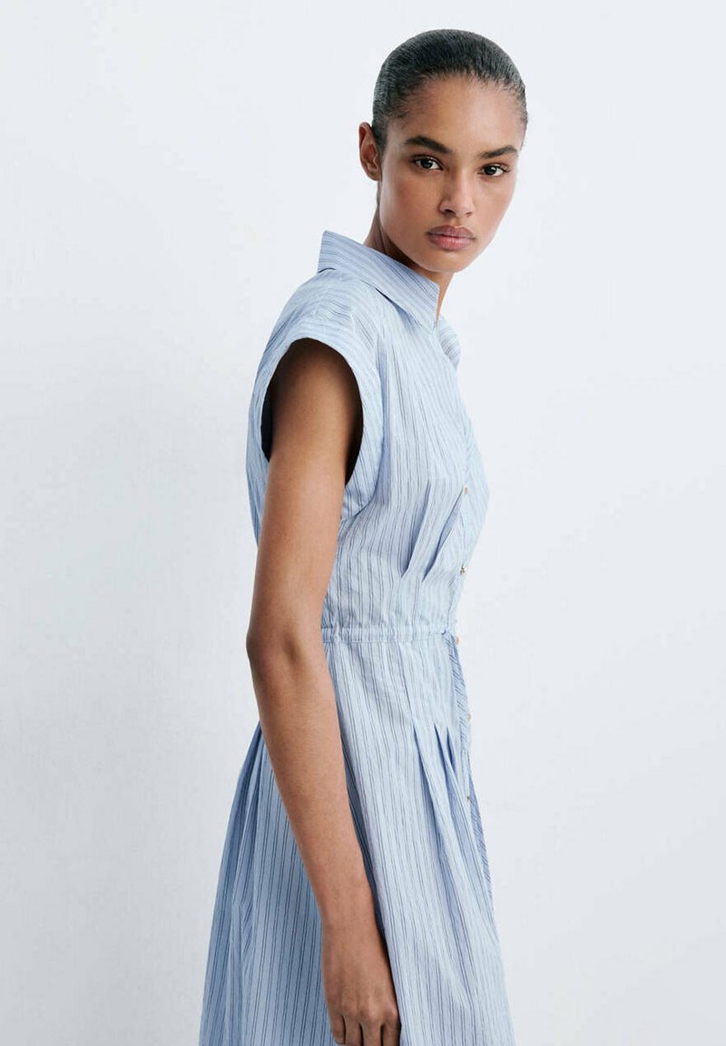 Jeune femme aux cheveux tirés en arrière, portant une robe sans manches rayée bleu clair, debout devant un fond blanc uni.