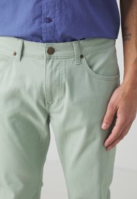 Pantalones de algodón verde claro con diseño de cinco bolsillos, herrajes de metal y costuras en contraste. Estilizados con una camisa morada ajustada.