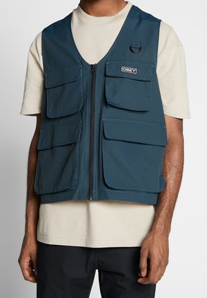 Man draagt een blauwe utility-vest met meerdere zakken over een beige T-shirt en zwarte broek, staat met ontspannen armen langs het lichaam.