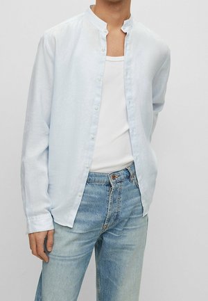 Homme portant une chemise bleu clair à col mandarin sur un débardeur blanc et un jean bleu clair, debout avec un bras derrière le dos.